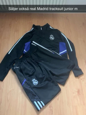 Real Madrid svart träningsset junior - Säljer ett svart träningsset från Real Madrid för juniorer, storlek M. Setet består av en långärmad tröja med dragkedja och matchande byxor. Båda plaggen har klubbens logga, vita och lila detaljer samt sportiga ränder på ärmar och ben.