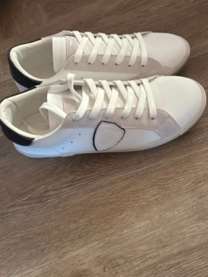Vita sneakers med svart detalj - Snygga vita sneakers med svart detalj på hälen och en vit patch på sidan. Skorna har vita snören och en stilren design med inslag av mocka och läder. Perfekta för dig som gillar en clean och modern look.