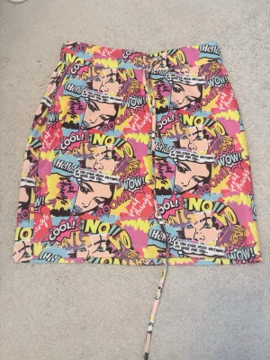 Färgglad pop art kortkjol med snörning - Säljer en unik kortkjol med färgglatt pop art-mönster i rosa, gul, blå och svart. Kjolen har snörning i midjan och coola serietidningsmotiv med ord som 'WOW', 'NO!' och 'COOL!'. Perfekt för dig som vill sticka ut med din stil.