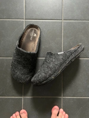 Mörkgrå ulltofflor från Shepherd - Mysiga mörkgrå tofflor från Shepherd i filtad ull med öppen häl och mjuk innersula. Klassisk slip-in modell med diskret logga på sidan och slitstark gummisula undertill. Perfekta för att hålla fötterna varma hemma.