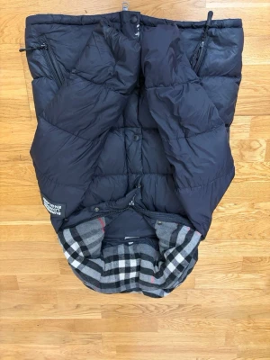 Burberry jacka - Snygg svart dunväst med rutigt foder i grått, vitt och rött. Västen har dragkedja, knappar och två framfickor med dragkedja. Märke på vänster sida med text. Perfekt för lager-på-lager och streetwear-stil. Finns lite som e sönder i mitten, Om du vill ha flera bilder skriv