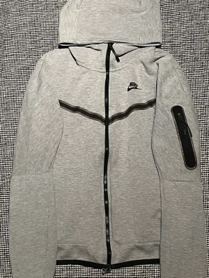 Grå Nike hoodie med dragkedja - Snygg grå hoodie från Nike med hel dragkedja framtill och svart Nike-logga på bröstet. Hoodien har huva, svarta detaljer och en praktisk ficka med dragkedja på ena ärmen. Perfekt för en sportig och avslappnad stil.