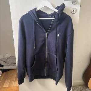 Ralph lauren zip hoodie  - Snygg marinblå hoodie från Polo Ralph Lauren med dragkedja framtill, två fickor och klassisk huva med dragsko. Ikoniska loggan broderad på bröstet. Perfekt för en avslappnad och stilren look.