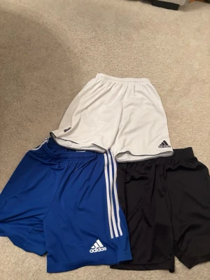 Adidas shorts - Tre par Adidas shorts i vitt, blått och svart. Klassisk sportig design med elastisk midja och Adidas-logga på vänster ben. De blå shortsen har vita ränder längs sidorna. Perfekta för träning eller sport. Säljer 3 för 189 kr