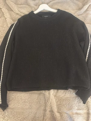 Svart stickad tröja Vero Moda - Mysig svart stickad tröja från Vero Moda med rund halsringning och vita detaljer längs ärmarna. Tröjan har en avslappnad passform och ribbad struktur som ger en cool och trendig look. Perfekt att matcha med jeans eller kjol.