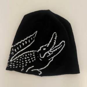 Svart mössa med vit krokodil - Säljer en svart mössa med en stor vit krokodil i grafiskt mönster på framsidan. Mössan är mjuk och stretchig, perfekt för kalla dagar. Passar dig som vill ha en cool och unik accessoar till din outfit.