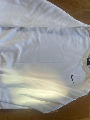 Vit sweatshirt från Nike - Klassisk vit sweatshirt från Nike med rund halsringning och diskret svart Nike-logga på bröstet. Tröjan har långa ärmar och ribbade muddar vid ärmslut och nederkant. Perfekt för en clean och sportig look.