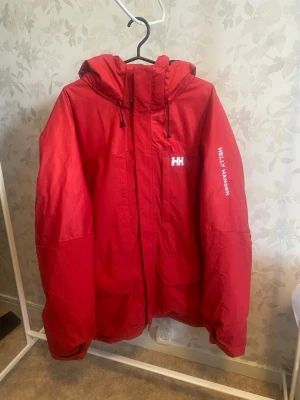 Helly Hansen Active Fall 2 Parka (Storlek XXL) - Snygg röd regnjacka från Helly Hansen med huva och dragsko. Jackan har två stora fickor framtill, dragkedja och tryckknappar. Helly Hansen-logga på bröstet och ärmen. Perfekt för regniga dagar och outdoor-äventyr.