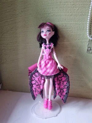 Monster High docka med rosa klänning - Säljer en Monster High docka med svart och rosa hår, klädd i en rosa klänning med svart mönster och detaljer. Dockan har stora rosa och svarta vingar med döskallemönster och rosa skor. Perfekt för samlare eller som present till någon som gillar Monster High.