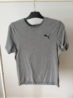 Grå t-shirt från Puma - En enkel och stilren grå t-shirt från Puma med klassisk rund hals och svart Puma-logga på bröstet. Tillverkad i mjuk bomull och har korta ärmar. Perfekt till vardags för en avslappnad look.