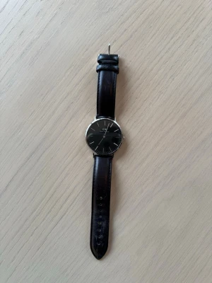 Svart klocka från Daniel Wellington - Stilren klocka från Daniel Wellington med svart urtavla och silverfärgat boett. Armbandet är i svart läder och har klassisk design. Perfekt accessoar för dig som. Levereras i originalask. Endast använd enstaka gånger.