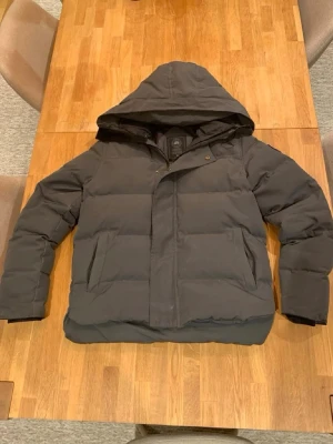 Canada goose macmillan - Säljer en mörkgrå pufferjacka med stor huva och dragkedja framtill. Jackan har två snedställda fickor och breda, vadderade paneler för extra värme. Perfekt för kalla vinterdagar och har en stilren, enkel design.