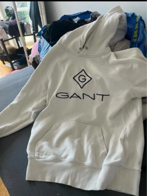 Vit hoodie från GANT med logga - Säljer en vit hoodie från GANT med stor broderad logga på bröstet. Hoodien har huva med dragsko och en klassisk magficka. Tillverkad i mjukt material och har ribbade muddar vid ärmslut och nederkant. Perfekt för en avslappnad och stilren look.