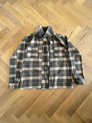 Rutig overshirt i grå och beige - Snygg overshirt i grått och beige rutmönster. Två bröstfickor med lock och knappar framtill. Rak passform och klassisk krage. Perfekt lager-på-lager-plagg för en avslappnad stil. Tillverkad i mjukt, vävt material.