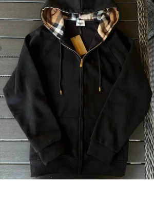 Svart hoodie med rutigt foder - Snygg svart hoodie med dragkedja och huva. Insidan av huvan har ett klassiskt beige, svart och vitt rutigt mönster. Tröjan har ribbade muddar vid ärmar och nederkant samt fickor framtill. Perfekt för en chill och stilren look.