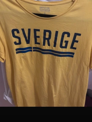 Gul Sverige t-shirt supporter - Gul t-shirt med blå texten 'Sverige' och två blå linjer under. Perfekt för att visa ditt stöd för landslaget. Rund halsringning och korta ärmar. Skön och enkel stil för alla tillfällen.