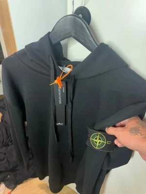 Svart hoodie från Stone Island - Säljer en svart hoodie från Stone Island med klassisk huva och dragsko. Hoodien har den ikoniska Stone Island-loggan på ärmen och en känguruficka framtill. Tillverkad i mjukt bomullsmaterial som är skönt att bära. Perfekt för dig som gillar streetwear och stilrena plagg.