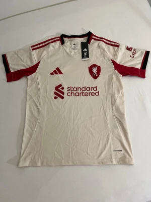 Liverpool bortatröja Adidas beige/röd - Snygg Liverpool FC bortatröja från Adidas i beige med röda detaljer och svarta inslag. Tröjan har klubbmärke och sponsortryck framtill samt röda ränder på axlarna. Tillverkad i lätt och ventilerande material, perfekt för fotboll eller träning.