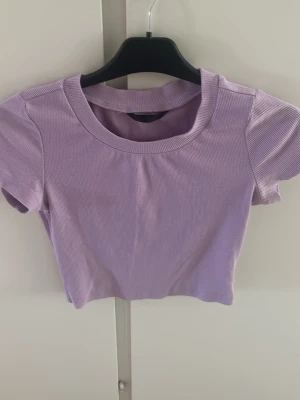 Ljuslila ribbad croppad topp - Säljer en ljuslila croppad topp med ribbad struktur och korta ärmar. Toppen har en rund halsringning och är perfekt för dig som gillar en enkel men trendig stil. Passar snyggt till både jeans och kjol.