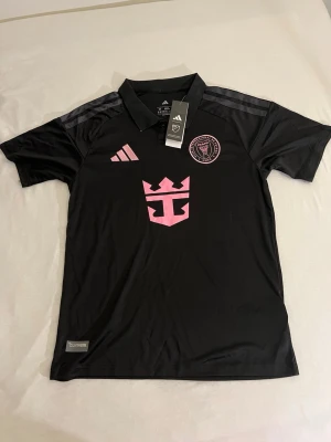 Svart Inter Miami matchtröja Adidas - Snygg svart matchtröja från Inter Miami med rosa detaljer och klubbmärke på bröstet. Tröjan har klassisk krage, korta ärmar och är tillverkad i ett lätt, ventilerande funktionsmaterial. Adidas-logga och CLIMACOOL-märke på framsidan.