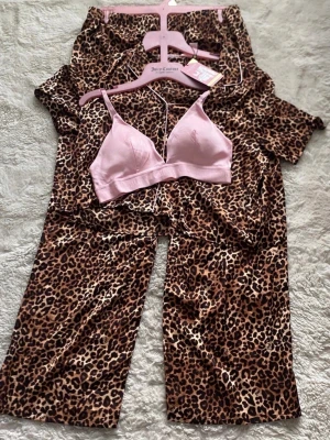 Leopardmönstrad pyjamas Juicy Couture size M - Snygg pyjamas från Juicy Couture med leopardmönster i brunt och svart. Setet består av kortärmad skjorta och byxor i satinliknande material samt en ljusrosa bralette med Juicy Couture-logga. Perfekt för mysiga hemmakvällar.