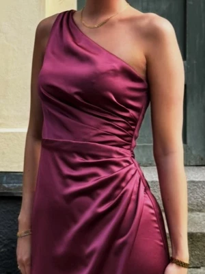 Vinröd oneshoulder långklänning - Snygg vinröd/ lila långklänning i glansigt material med oneshoulder-design och draperad midja. Klänningene har en elegant slits på sidan och smickrande passform som framhäver figuren. Perfekt för speciella tillfällen. Jag hade den en kväll på min bal OBS JAG ÄR 1,78. nypris va 4500kr
