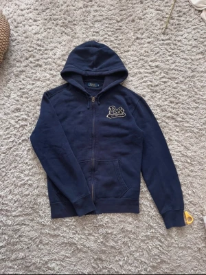 Mörkblå hoodie med dragkedja Polo - Mörkblå hoodie från Polo med dragkedja framtill och broderad logga på bröstet där det står 'Polo EST 1967'. Hoodien har huva, två fickor och ribbade muddar vid ärmar och nederkant. Perfekt för en avslappnad och stilren look.