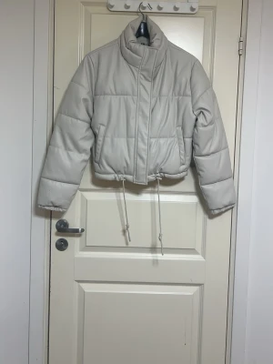 Ljusgrå pufferjacka med dragsko - Snygg ljusgrå/ vit  pufferjacka med hög krage och dragsko i midjan. Jackan har en kortare, boxig passform och stängs med dragkedja och tryckknappar. Perfekt för kalla vinterdagar och ger en trendig look. Har aldriga använt den, prislappen finns kvar. Mycket snygg jacka.