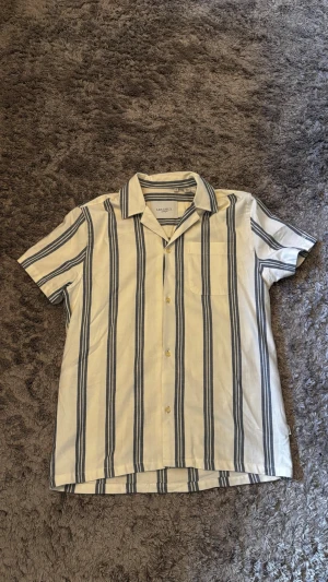 Randig skjorta från Les Deux XS - Kortärmad skjorta från Les Deux i vitt med breda blå ränder. Skjortan har klassisk krage, bröstficka och knappar framtill. Tillverkad i ett mjukt bomullsmaterial som passar perfekt till sommaren.