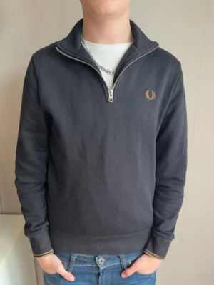 Svart half-zip tröja Fred Perry - Svart långärmad tröja från Fred Perry med half-zip och ståkrage. Tröjan har diskret logga broderad i guld på bröstet och ribbade muddar vid ärmslut och nederkant. Perfekt till jeans för en clean och stilren look.
