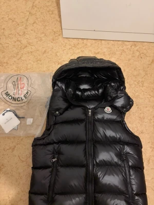 Moncler svart dunväst - Snygg svart dunväst från Moncler med glansig finish och avtagbar huva. Västen har två dragkedjeförsedda fickor framtill och klassisk Moncler-logga på bröstet. Perfekt för lager-på-lager och håller dig varm med stil. Med nfc Scan och taggar som kommer med. Storlek S men liten i storleken så sitter också som en XS typ. Och som är perfekt för detta väder som kommer.