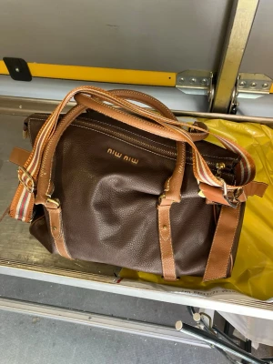 Brun handväska  - Snygg brun handväska i mjukt ”läder” med detaljer i ljusbrunt och guldfärgade spännen. Väskan har både handtag och en randig axelrem för extra stilpoäng. Perfekt för dig som vill ha en trendig och rymlig väska.