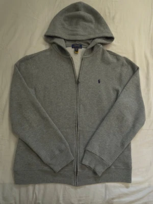 Grå zip hoodie från polo Ralph Lauren - Snygg grå hoodie med dragkedja och huva. Klassisk design med två stora fickor framtill och diskret logga på bröstet. Perfekt till jeans och enkel att matcha med andra plagg. Barn xl 18-20 men passar en mindre S