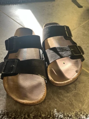 Svarta Birkenstock sandaler mocka - Säljer ett par klassiska svarta Birkenstock sandaler i mocka med två breda remmar och justerbara metallspännen. Fotbädden är i kork och innersulan är mjuk och formad för extra komfort. Perfekta för en avslappnad och stilren look.
