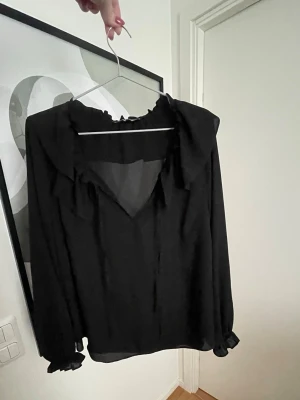 Svart blus  - Svart blus i ett tunt, transparent tyg med volangdetaljer vid kragen och ärmsluten. Från Primark i storlek L