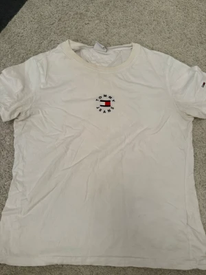 Vit T-shirt från Tommy Jeans - Vit T-shirt från Tommy Jeans med rund hals och klassisk logga broderad på bröstet. T-shirten har korta ärmar och är gjord i mjuk bomull, perfekt för en avslappnad och stilren look. 