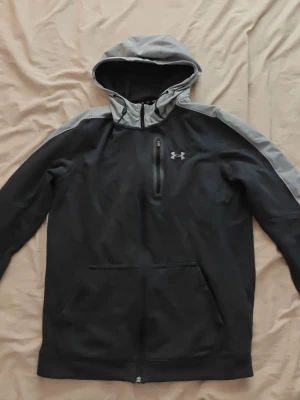 Under Armour Zip Hoodie - Svart hoodie från Under Armour med grå huva och grå detaljer längs axlarna. Tröjan har dragkedja vid halsen, en bröstficka med dragkedja och en stor magficka. Perfekt för en sportig och avslappnad stil.