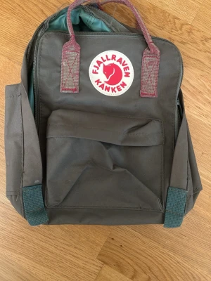 Grön Fjällräven Kånken ryggsäck - Klassisk grön Fjällräven Kånken ryggsäck med vinröda handtag och axelband. Ryggsäcken har en stor ytterficka med dragkedja, två sidofickor och det ikoniska runda Kånken-märket framtill. Tillverkad i slitstarkt syntetmaterial. Bleknade handtag (se bild)  
