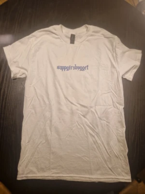 Vit t-shirt med tryck från Gildan - Vit t-shirt från Gildan med blå texten 'soppairobrygget' tryckt på bröstet. Klassisk rak passform och rund halsringning. Tillverkad i bomull för en mjuk och bekväm känsla. Perfekt för dig som gillar enkel och stilren design.