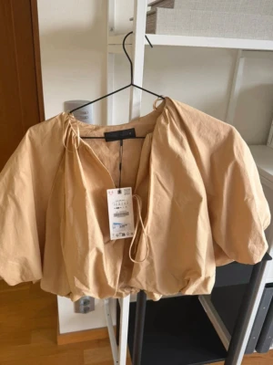 Beige croppad topp med puffärm - Trendig beige croppad topp från ZARA med voluminösa puffärmar och knytning framtill. Tillverkad i ett lätt bomullsliknande material som ger en luftig känsla. Perfekt för att skapa en cool och avslappnad look.