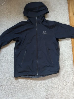 Mörkblå regnjacka från Arc'teryx - Snygg Mörkblå regnjacka från Arc'teryx med huva och hel dragkedja framtill. Jackan har två snedställda fickor med dragkedja och diskret logga på bröstet. Tillverkad i ett vattentätt material som passar perfekt för regniga dagar. Hör av er vid funderingar och gärna innan ni köper!