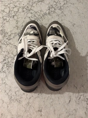 Camo sneakers med vita detaljer - Snygga sneakers med grå, svart och vit camo-mönster och vita snören. Skorna har svarta och grå detaljer samt svart innersula. Perfekta för dig som vill sticka ut med en cool streetwear-look. Låg modell som passar till många olika outfits.