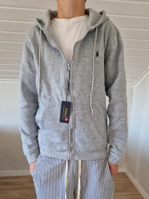 Polo Ralph Lauren zip hoodie - Snygg grå hoodie från Polo Ralph Lauren med dragkedja framtill, huva med dragsko och diskret logga på bröstet. Tillverkad i mjukt sweatshirtmaterial som passar perfekt till chill eller streetwear. Två fickor framtill för extra casual vibe.