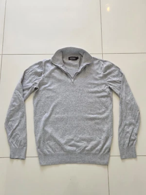 Ljusgrå 1/4 zip tröja från Dressmann - Ljusgrå stickad tröja från Dressmann med halvzip och hög krage. Tröjan har långa ärmar och ribbade muddar vid ärmslut och nederkant. Enkel att matcha och perfekt för lager på lager.