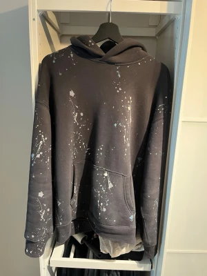 Svart hoodie med färgstänk Abercrombie - Säljer en svart hoodie från Abercrombie & Fitch i mjuk fleece med unikt färgstänksmönster i vitt och ljusblått. Klassisk känguruficka framtill och stor huva. Perfekt för dig som gillar streetwear och vill sticka ut lite extra.