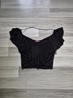 Svart croppad topp med volang - Snygg svart croppad topp med bred volang runt hela ringningen och offshoulder-design. Perfekt för dig som vill ha en trendig och luftig topp till sommaren. Tillverkad i mjukt material som känns skönt mot huden.