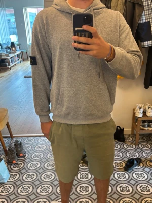 Grå hoodie från Stone Island - Snygg grå hoodie från Stone Island med klassisk patch på vänster ärm. Hoodien har dragsko i huvan och en stor magficka. Tillverkad i mjukt material som känns skönt mot huden. Perfekt för en avslappnad och stilren look.
