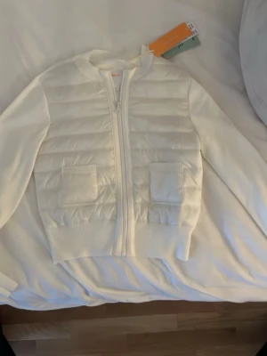 Vit quiltad cardiganjacka med fickor - Snygg vit cardiganjacka med quiltade partier framtill och två praktiska fickor. Dragkedja hela vägen och ribbade muddar vid ärmslut och nederkant. Perfekt för lager på lager och ger en clean look till din outfit. Helt ny med prislapp,aldrig använd💕