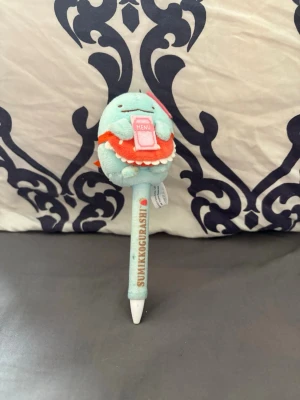 Sumikkogurashi blå bläckpenna - Söt bläckpenna med Sumikkogurashi-figur i blått plyschmaterial på toppen. Figuren håller en rosa meny och har en orange förkläde. Pennan har text och små dekorationer längs skaftet. Perfekt för dig som gillar kawaii och japanska karaktärer.