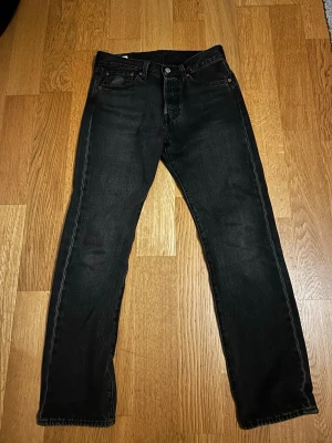 Svarta Levi's 501 jeans W28 L30 - Klassiska svarta Levi's 501 jeans med rak passform och knappgylf. Jeansen är tillverkade i slitstark denim och har fem fickor samt den ikoniska läderpatchen bak i midjan. Perfekta för en tidlös och avslappnad stil.